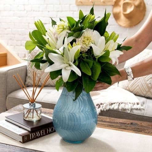 large blue ean. cremation urn for human ashes that looks like flower vase. Ladies hands placing the funeral urn on coffee table. Urn has white lillies displayed. woman wears silver bracelet. White background with sofa, wood stairs, books and diffuser.