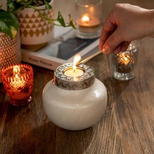 Small candle urn for human ashes or pets displayed on wooden table.. keepsake urn is white with engraved silver lid. Ladies hand is lighting a candle in the top of the ashes urn with a match. small tealight candles and plants in background on table.