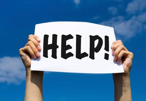 Ladies hand holding up HELP! sign with Help printed in black on white paper. Blue sky and cloud background. Bright image.
