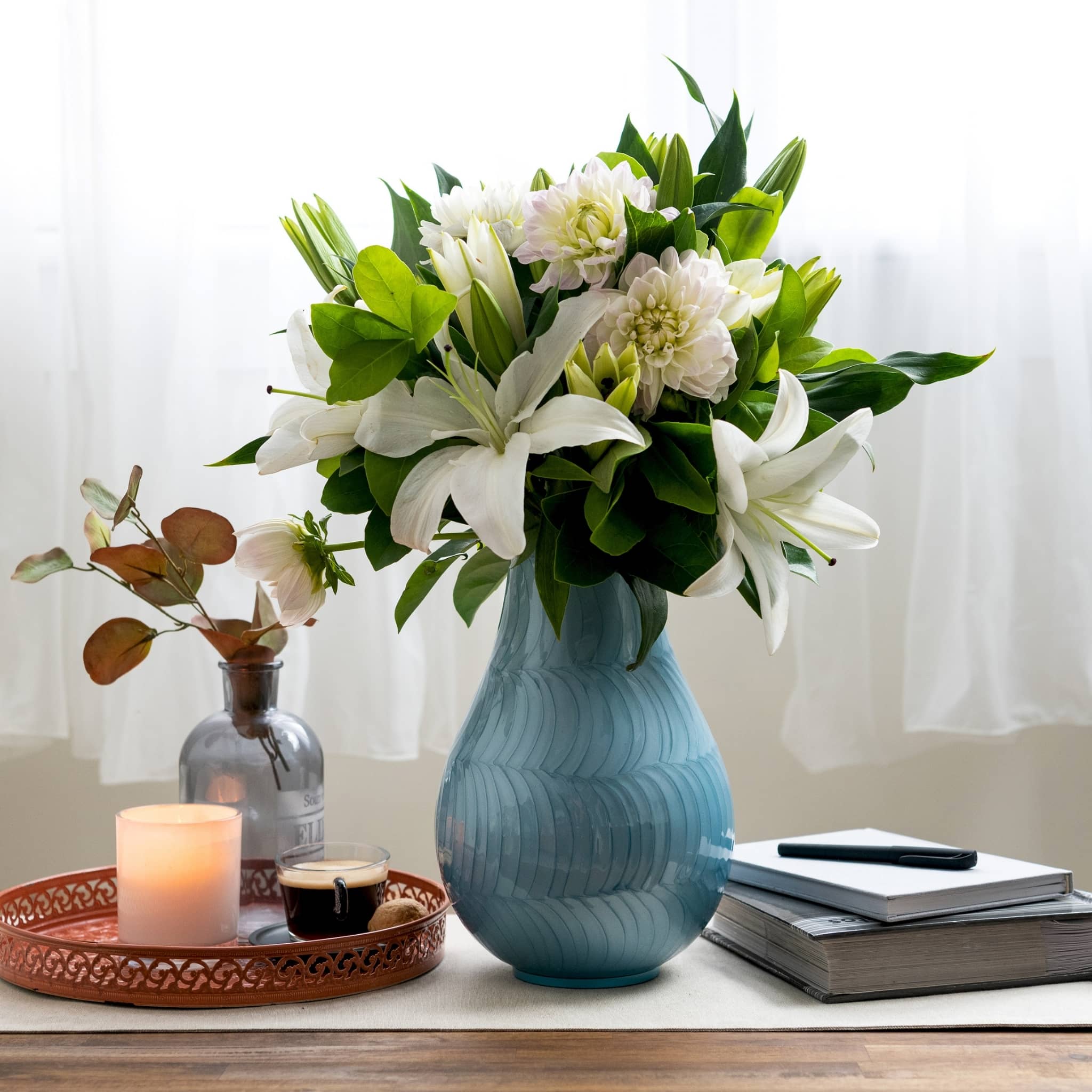blue cremation vase for ashes. ash vase displayed on table in front of window and white curtain. Displays floral bouquet of white and green.  books, candle, and coffee displayed on tabletop