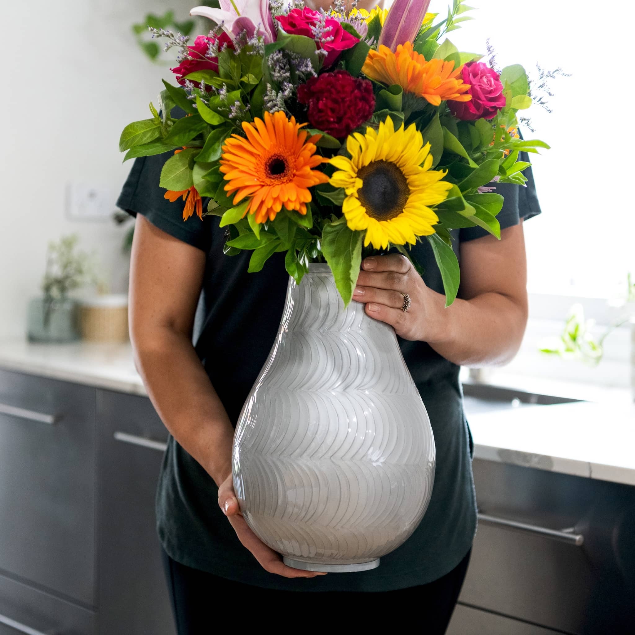 woman in black holds large ash urn for cremated remains in hands infront of ladies waist. flower ashes vase has bright floral display. shot in kitchen. picture depicts extr LARGE URN SIZE.