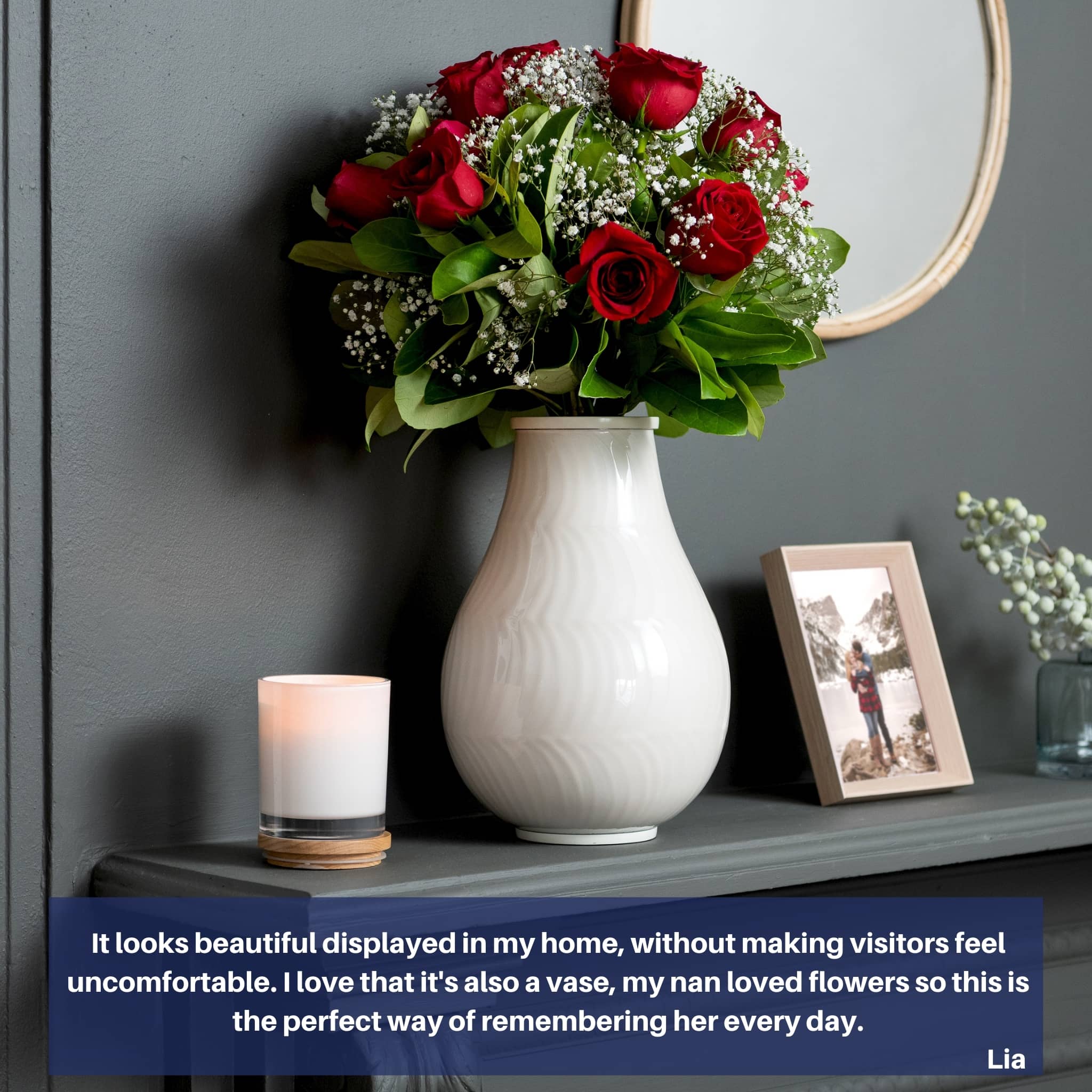 large cremation urn for human ashes adult is displayed on mantle with red rosed inside. candle and phioto sit either side of this large whitehuman ash urn.  dark gray background wall with mirror. red roses, green leaves and small white flowers.