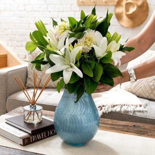 large blue ean. cremation urn for human ashes that looks like flower vase. Ladies hands placing the funeral urn on coffee table. Urn has white lillies displayed. woman wears silver bracelet. White background with sofa, wood stairs, books and diffuser. 