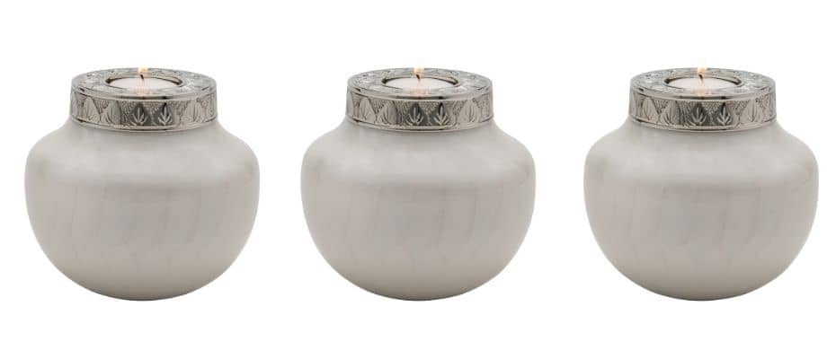 three small  mini tea light urns urns in a line. White keepsake urns for human ashes or pets. White finish makes them appear ceramic even though they are metal. Each min urn has a silver lid with engraving. Displayed with a lit candle in the top.