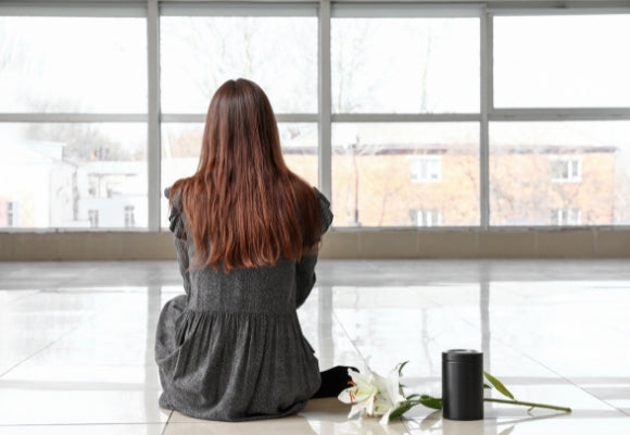  Young female sat on glossy white floor looking out the glass window. Lady is wearing gray and sat beside a small black traditional keepsake urn for human ashes and  a white funeral lily.