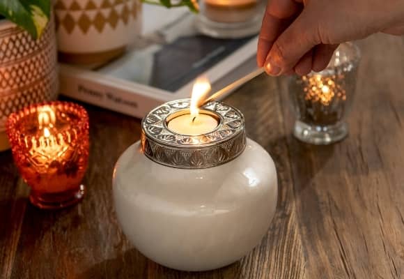Small keepsake urn for human ashes and pets. White mini urn with engraved silver lid. Canlde is being lit with a match in womans hand. Mini urn for human ashes is displayed on wooden table top with book, plant and small candles. Pot plant has gold pattern