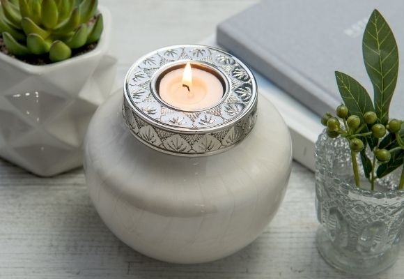 Mini urn. white mother-of-pearl tealight urn. Candle cremation urn is central, keepsake urn for ashes on table with small plants and notebooks. Candle lit in tea-light holder on keepsake urns silver lid.  Gray notebook with silver writing in background. 