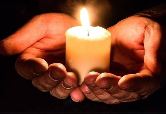 hands holding a pillar candle. Candle is lit with bright yellow flame. Dark background. 