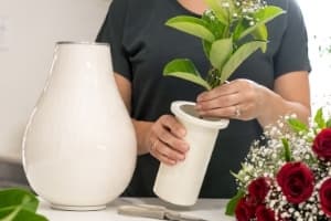 White Memorial Vase Urn for human ashes. Roses being put into ash vase insert by woman. Earn is white, looks ceramic but is metal. Large size displayed on side as lady arranges flowers. More flowers & scissors on side. White floral urn. Flower vase urn. 